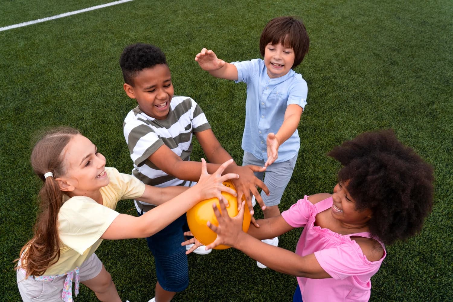 Four children reaching for an orange ball together outdoors on a grass field